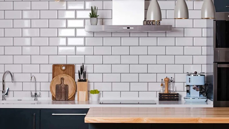Стена на модерна кухня, облицована с бели гланцирани плочки тип „метро“ с тъмна фуга над светъл дървен плот.