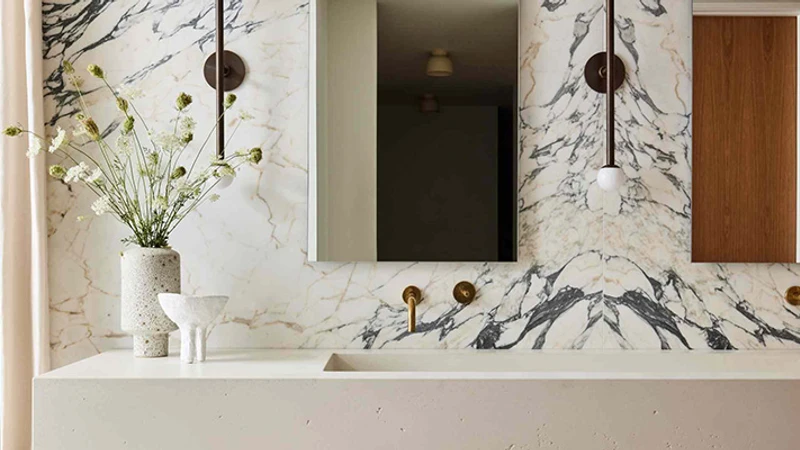 Луксозна баня с голямо-форматни плочки от бял мрамор със сиви жилки, съчетани с минималистична мивка и златни батерии.
